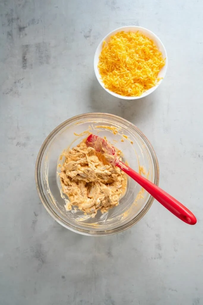 Combining shredded chicken with buffalo sauce in a bowl. A bowl of grated cheese next to it.