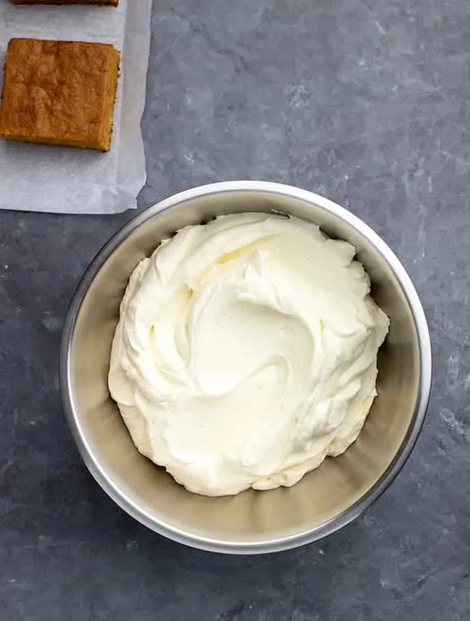 Cream cheese frosting in a large round metal bowl with plain pumpkin square in view