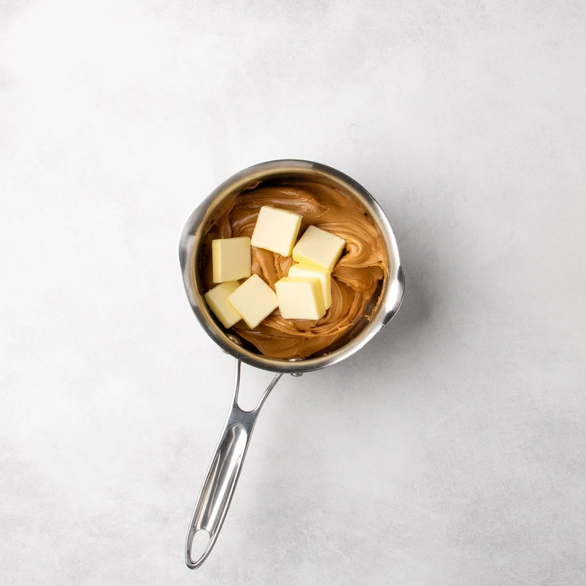 Cubes of butter placed on top of peanut butter in a small metal saucepan on a light surface.