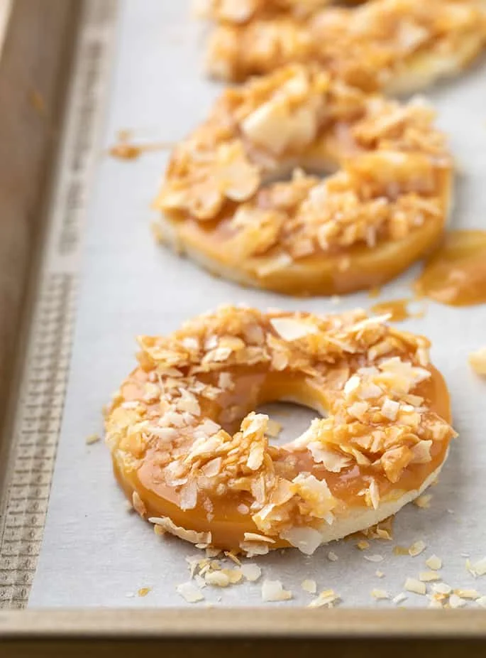Donut shaped cookies coated in caramel and crushed toasted coconut on white paper on baking tray