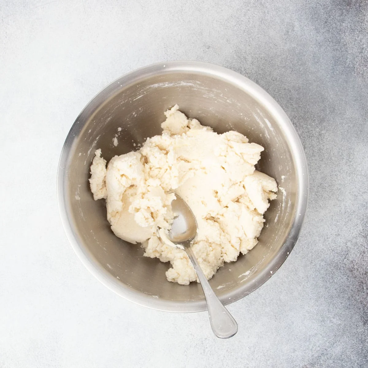 Dough fully mixed and rough in texture in a metal bowl with spoon.