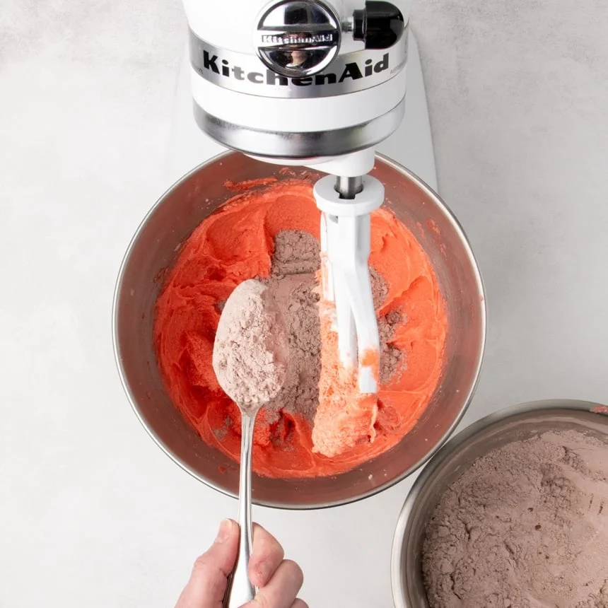 Dry ingredients being added to red batter in a stand mixer bowl using a spoon.