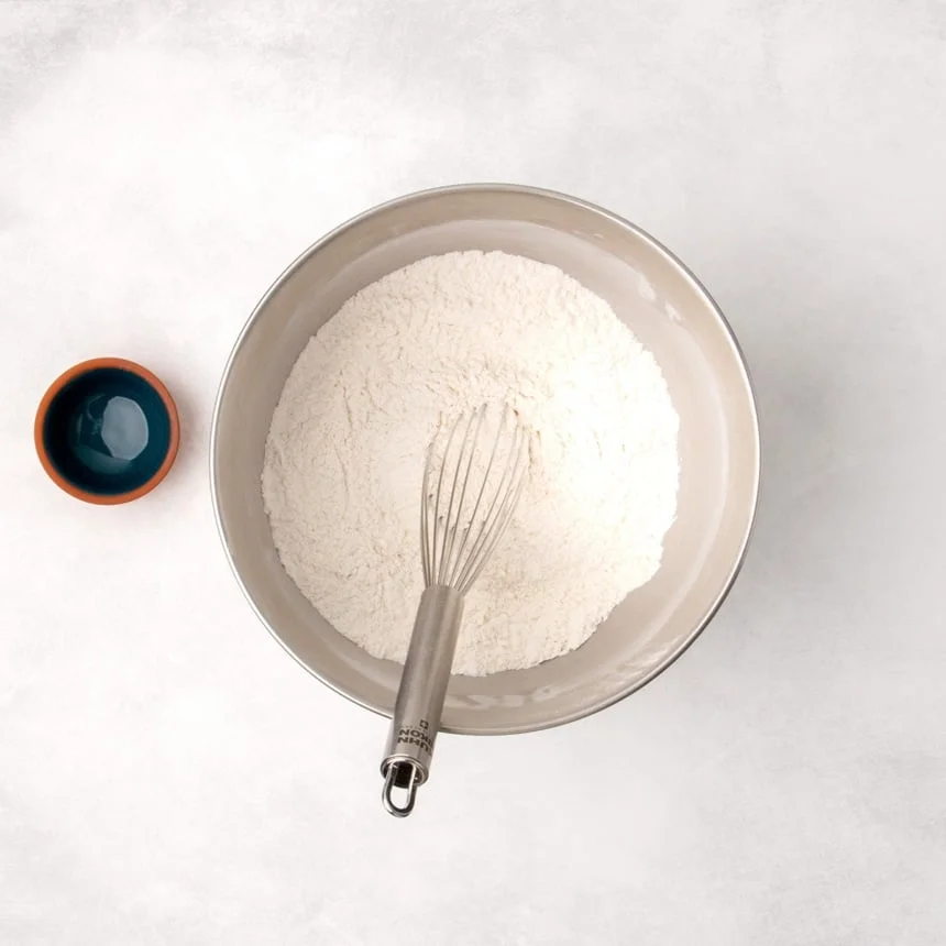 Dry ingredients mixed together in a metal bowl with a whisk resting inside, next to a small empty bowl.