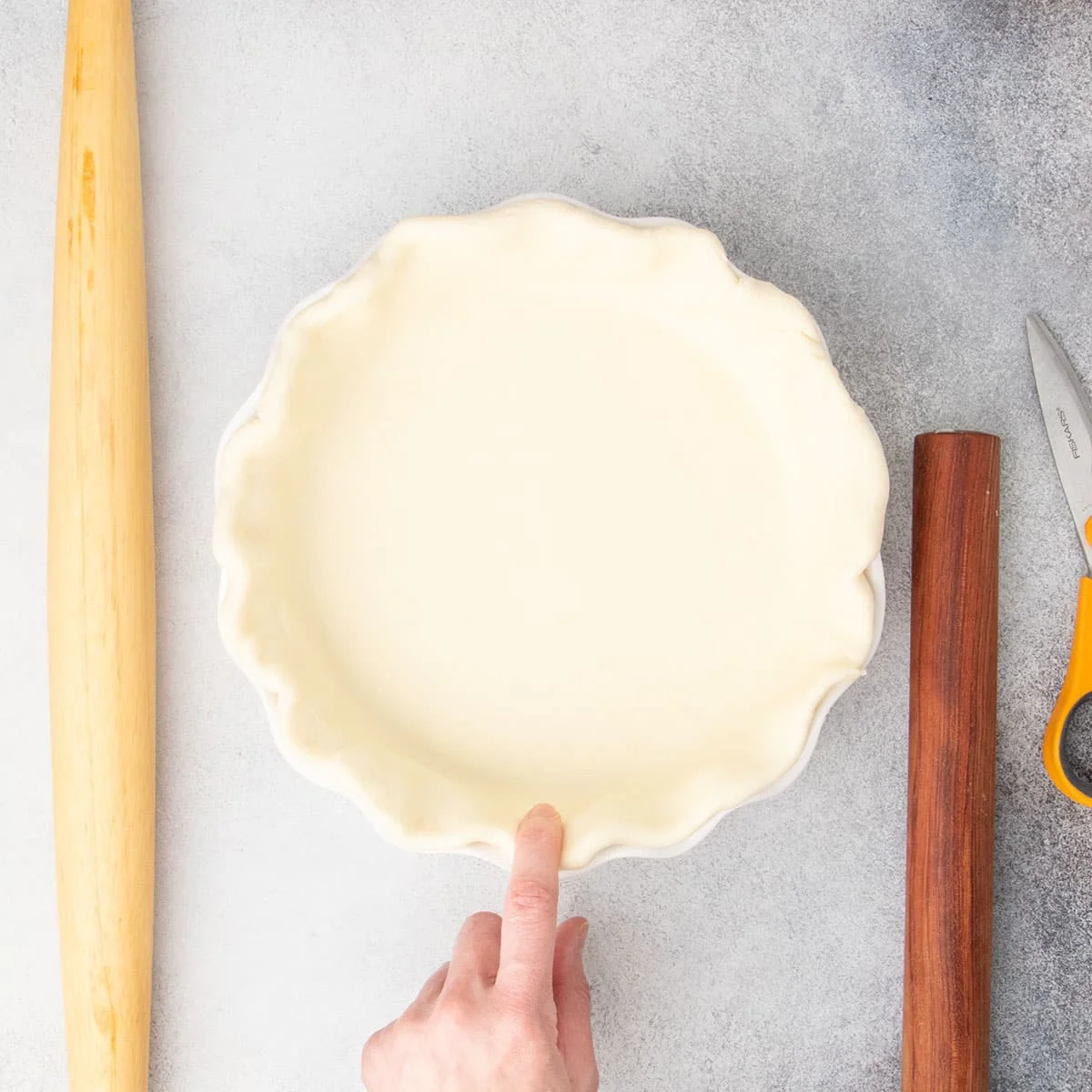 Finger pressing edge of shaped pie raw pie crust in white pie dish with two rolling pins and kitchen shears nearby on light gray surface.