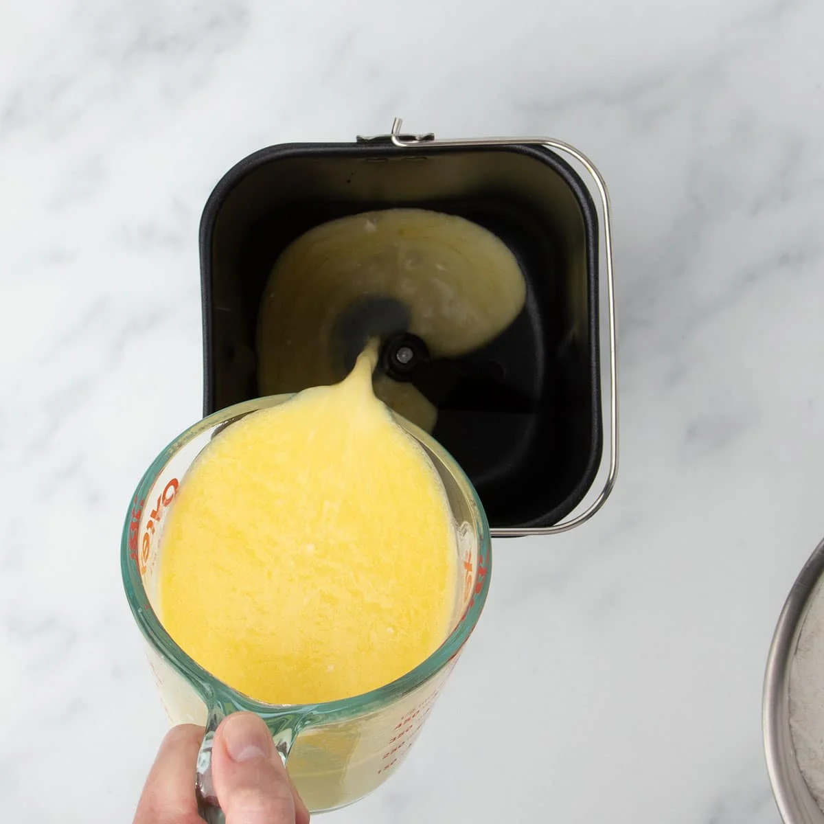 fingers holding 2 cup glass measuring cup pouring yellow mixture into baking pan for bread machine