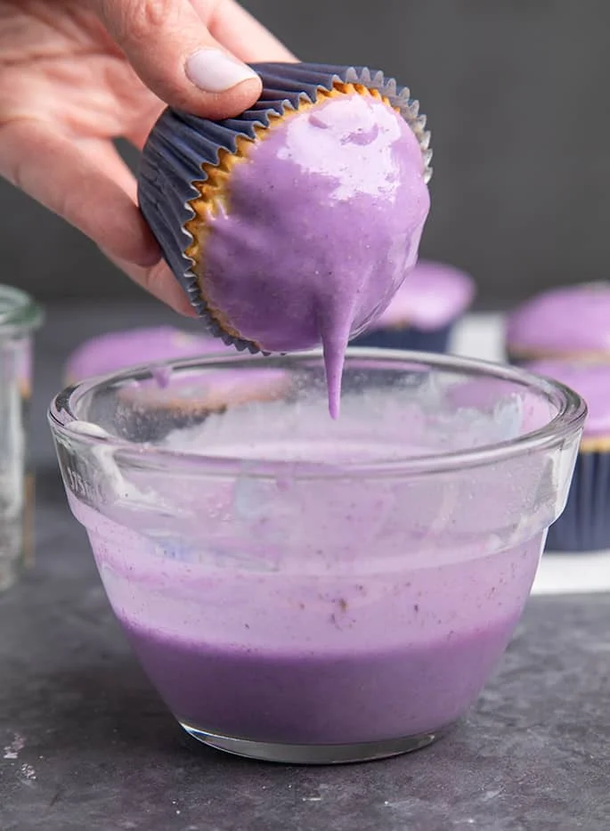 Fingers holding cupcake with dripping lavender glaze over glass bowl.