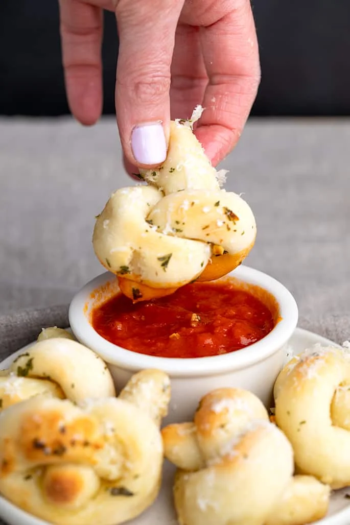 Fingers holding garlic knot that's been dipped in tomato sauce
