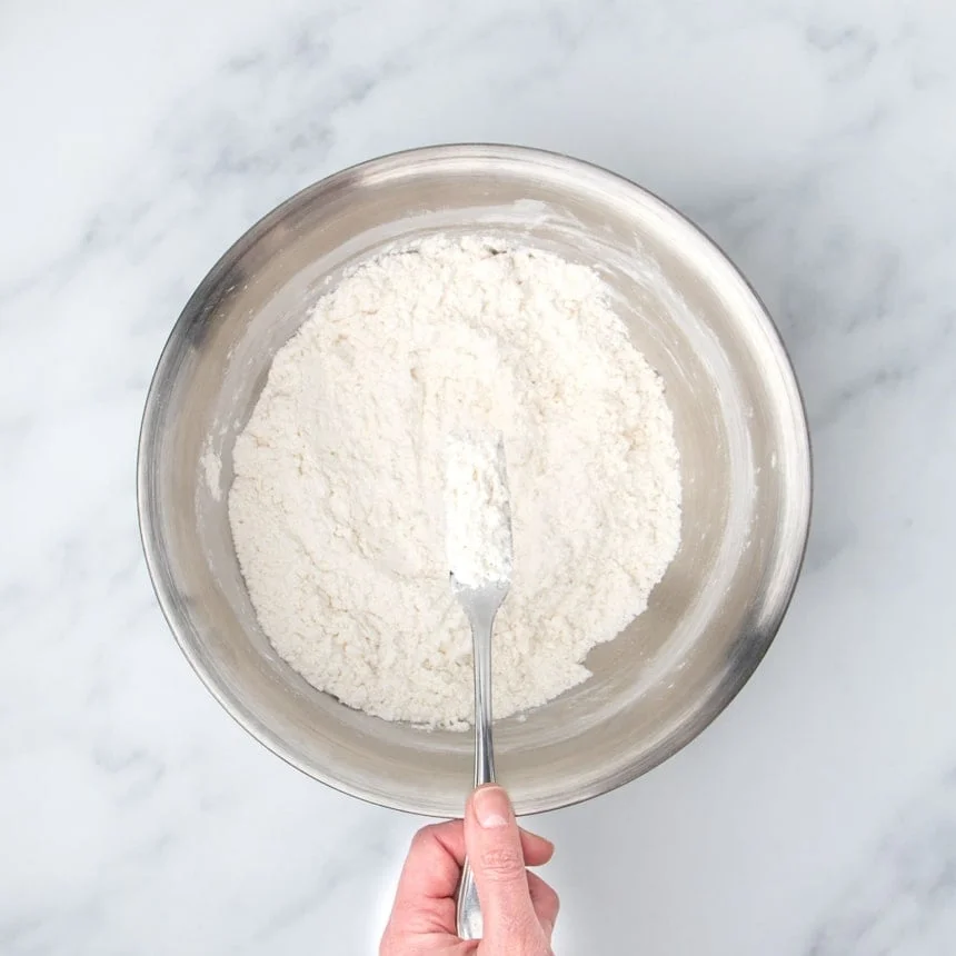 fingers holding metal fork with sandy white mixture of coconut oil mixed into flour, held over same mixing bowl