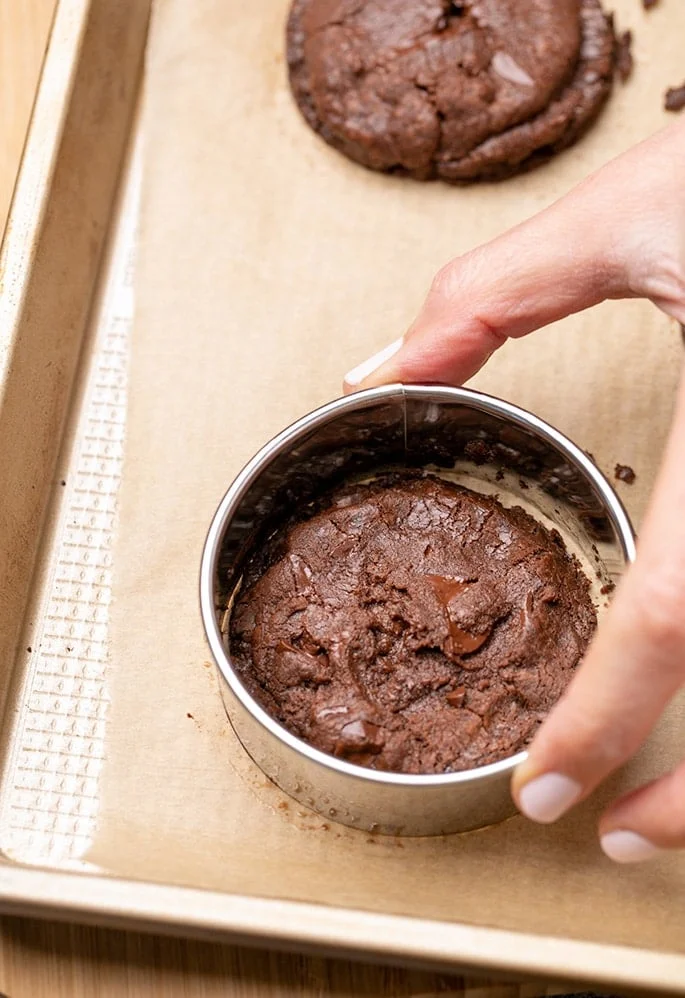 Fingers holding round cookie cutter around brown cookie on baking sheet