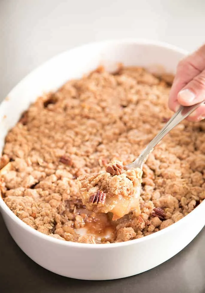 Fingers holding silver serving spoon with a spoonful of cooked sliced apples, brown crumb topping and pecans above a white casserole dish