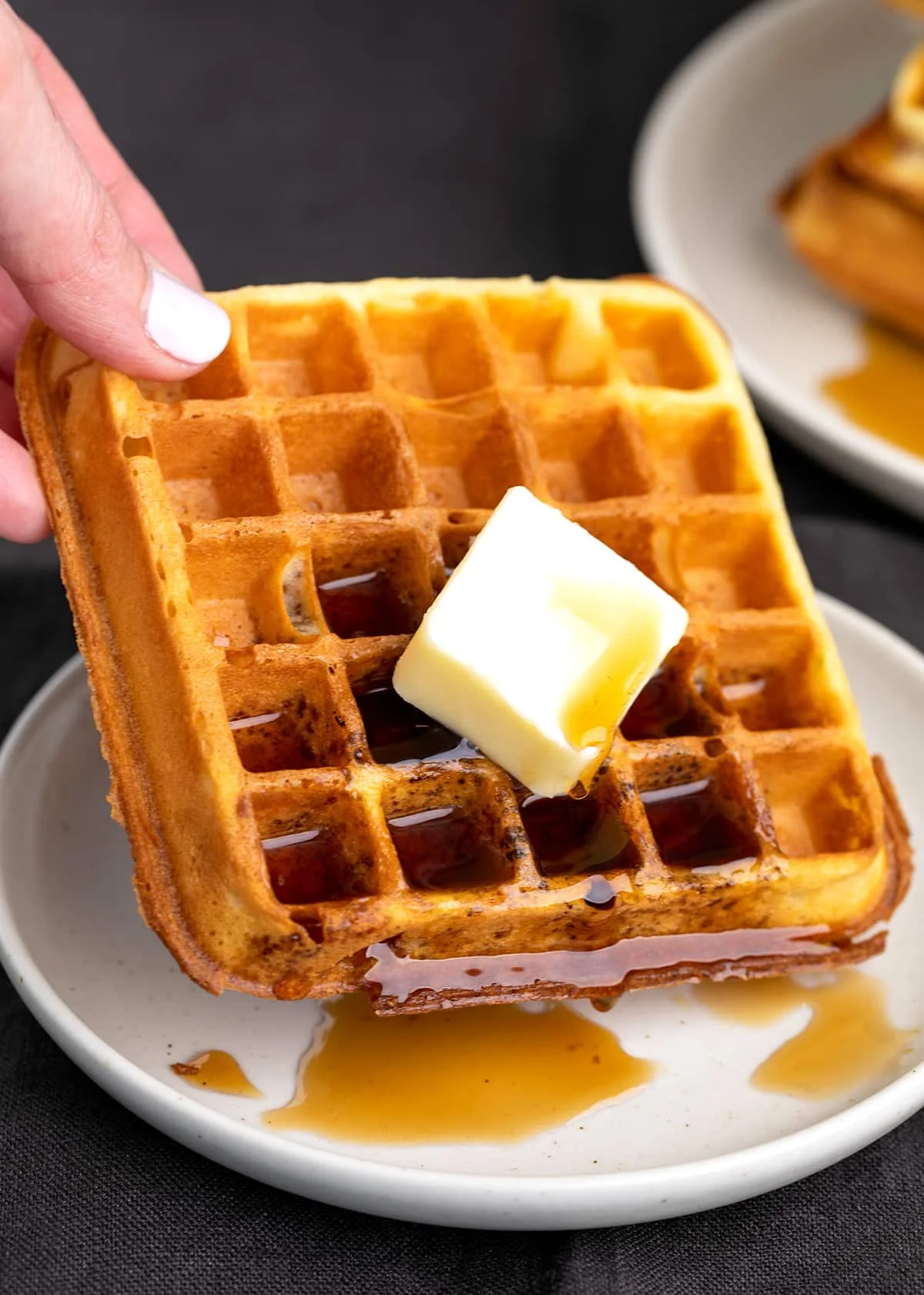 Fingers holding waffle with syrup and butter dripping on small plate