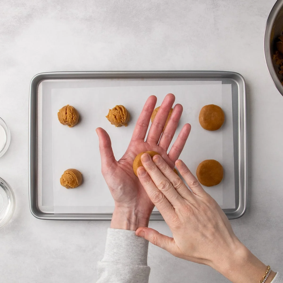 Fingers of one hand pressing a ball of peanut butter cookie dough flat in the palm of another hand, held over a baking sheet with other dough portions.