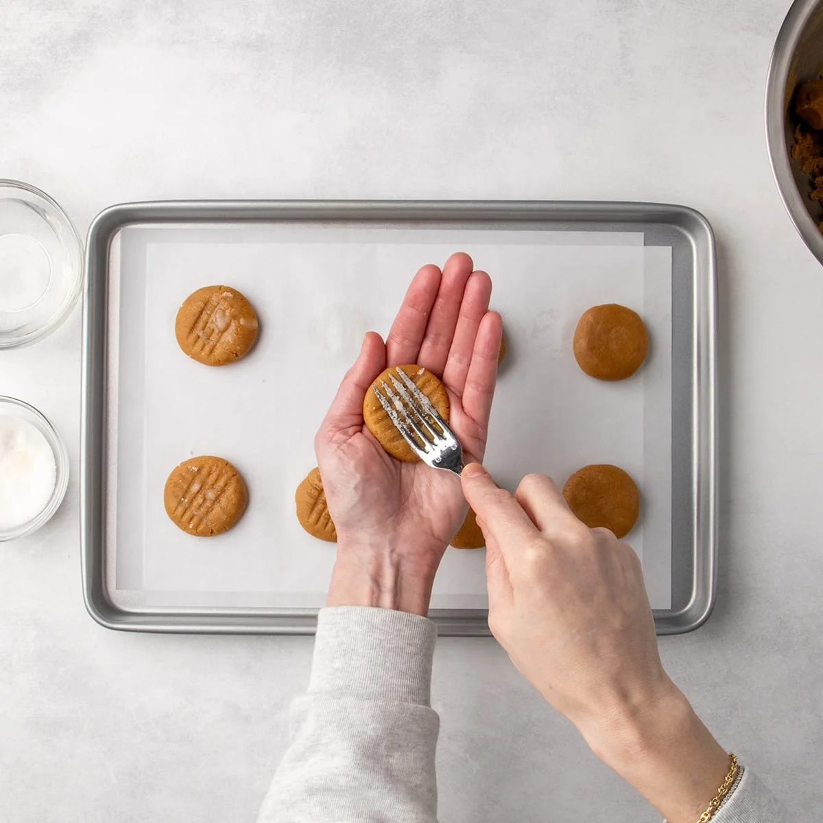 Fingers of one hand pressing a fork into a peanut butter cookie dough disk to create crosshatch marks over a parchment-lined baking sheet.