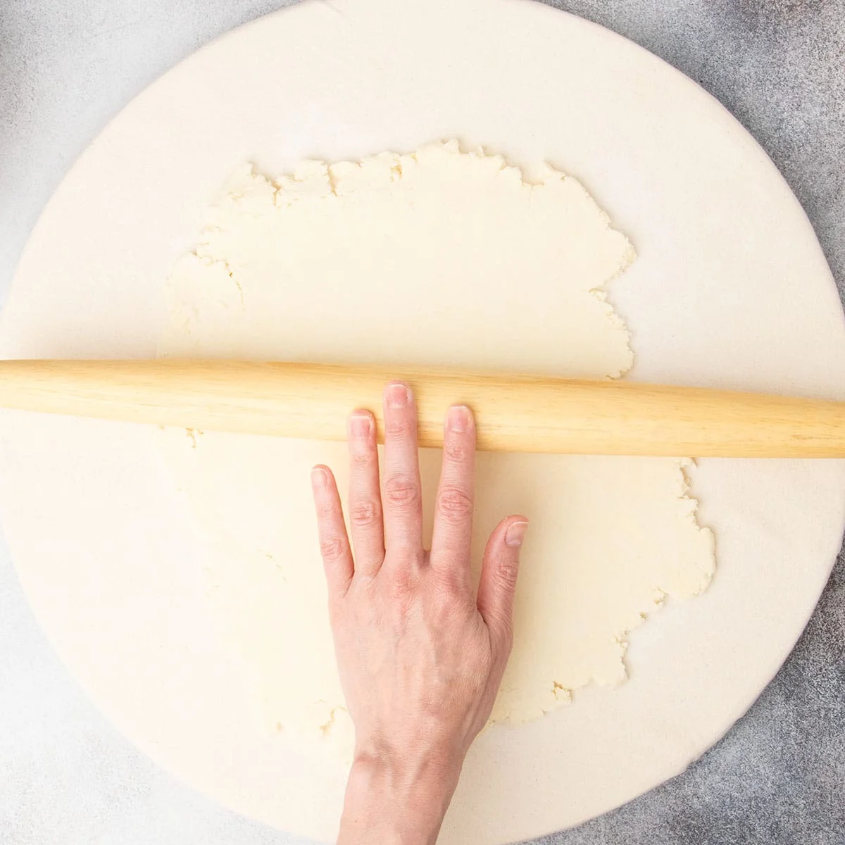 Fingers rolling blonde wood tapered rolling pin to roll out light tan pie crust dough into rough rectangle on canvas covered pastry board.