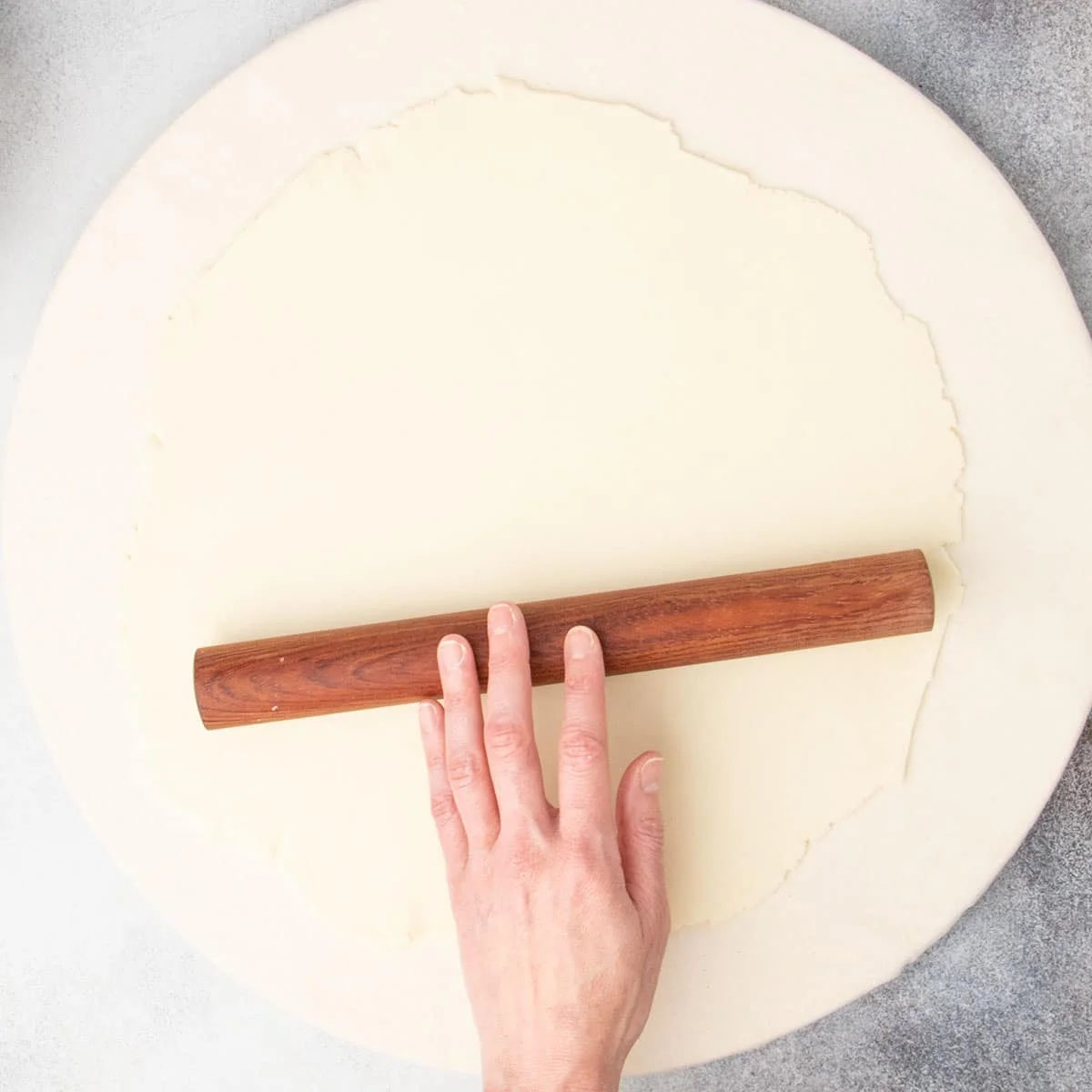 Fingers rolling short dark wood rolling pin on 10-inch round raw pie crust on board.