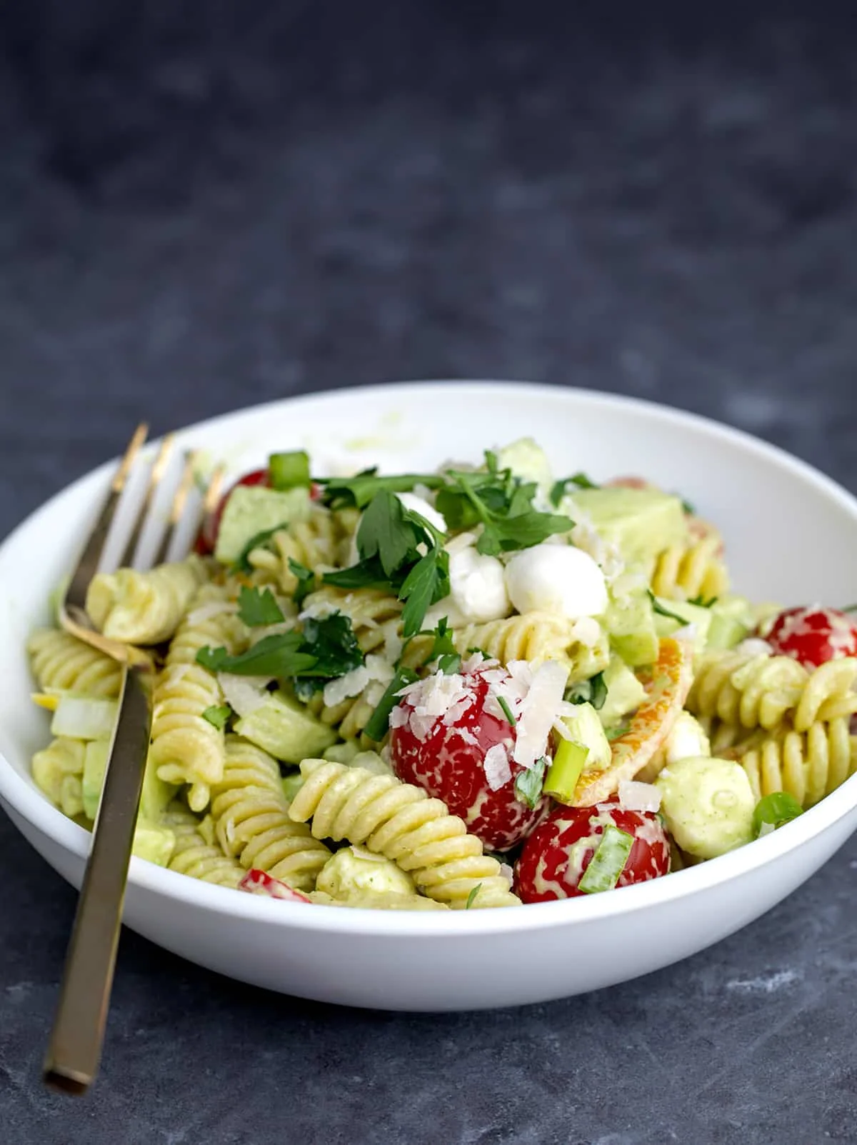 flat white bowl with light green colored dressing on cooked fusilli pasta, tomatoes, parsley, shaved cheese, white cheese pearls with a metal fork on a gray surface
