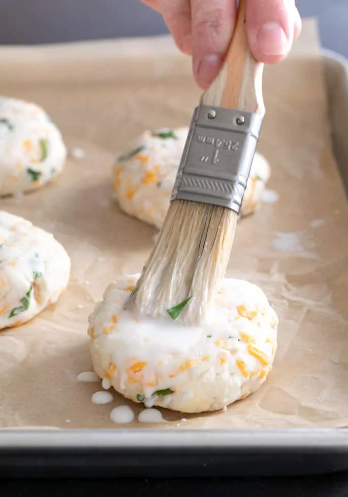 Flattened gluten free cheddar bay biscuits being brushed with cream
