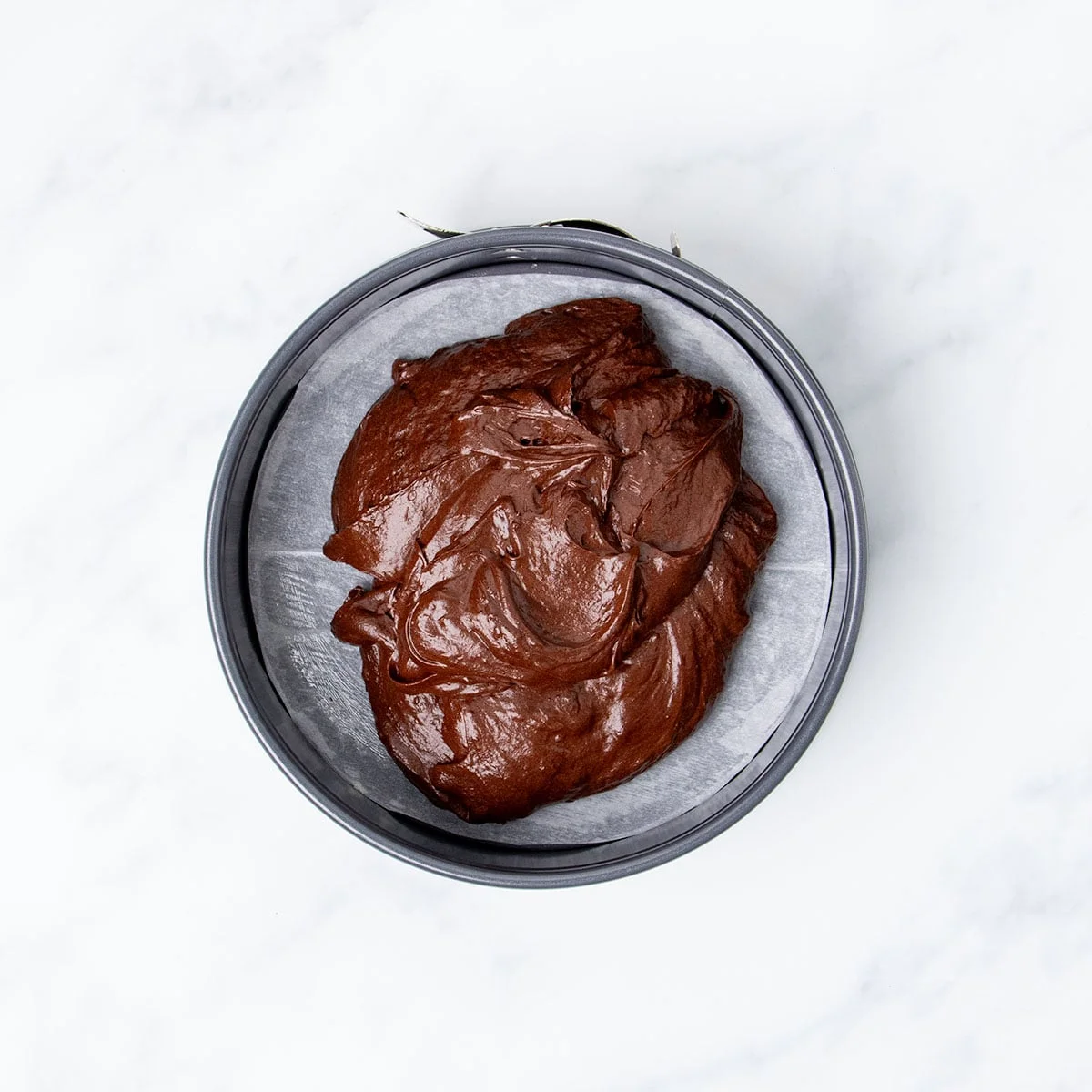Fluffy brown flourless chocolate cake mixture in round gray springform cake pan lined in white round of paper.