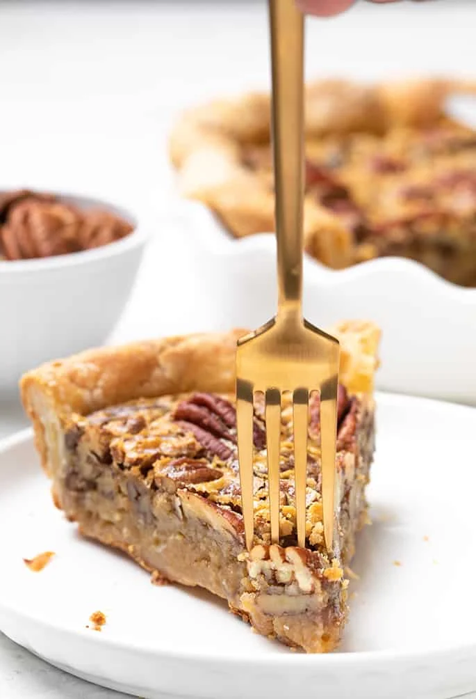Fork being inserted into slice of gf pecan pie on small white plate with small bowl of pecans and rest of pie in background