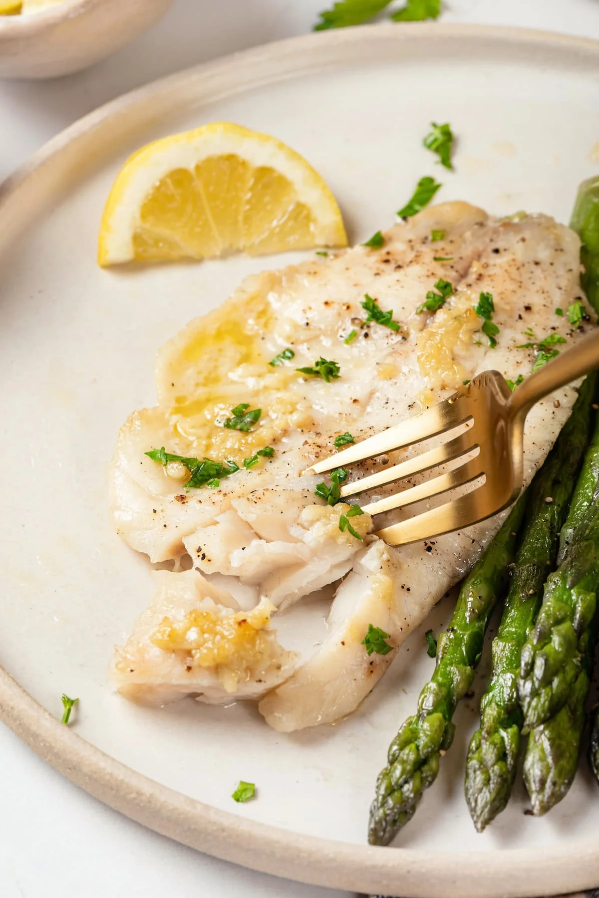 Fork cutting into baked tilapia on plate with asparagus