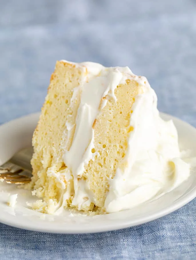 fork on side of slice of 2 layer lemon cake with lots of white frosting on small white plate on blue cloth