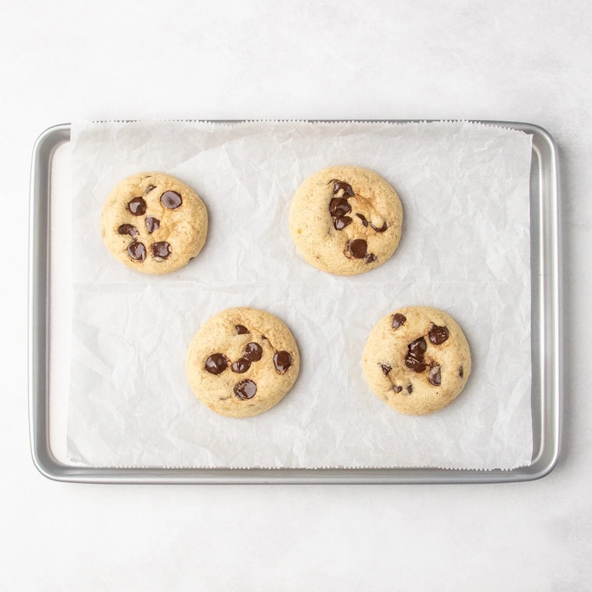 Four baked pale chocolate chip cookies on a parchment-lined baking sheet.