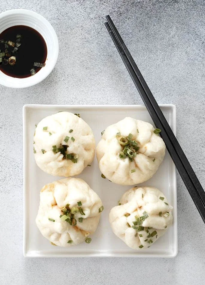 Four Chinese meat buns on a plate with chop sticks and soy sauce in a small bowl