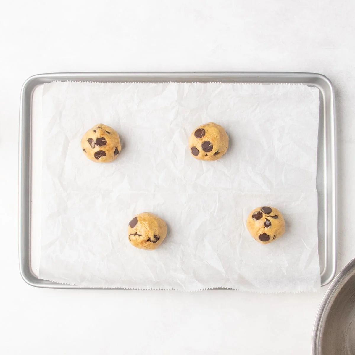 Four rounded balls of chocolate chip cookie dough on a parchment-lined baking sheet.