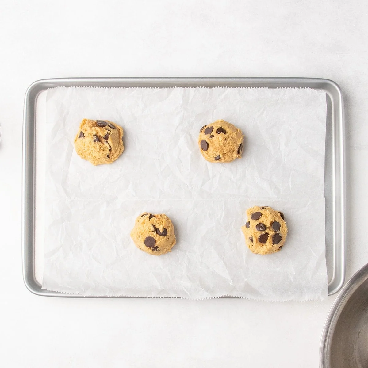 Four scoops of chocolate chip cookie dough on a parchment-lined baking sheet.
