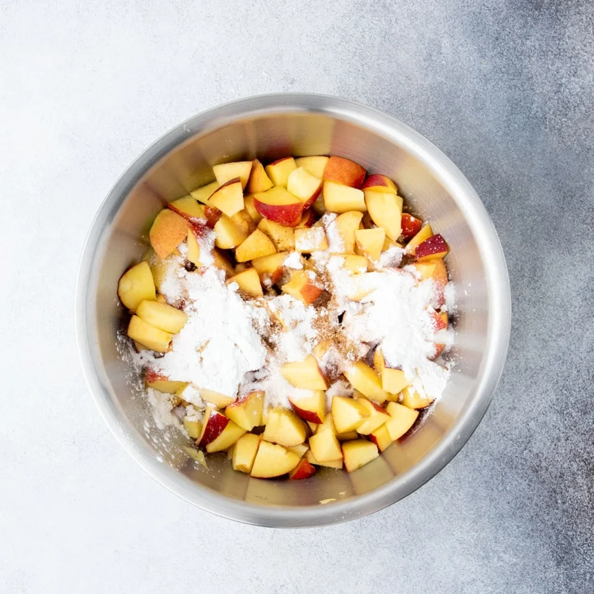 Fresh peach chunks in a large metal bowl topped with starch, sugar, and spices before mixing for the filling.