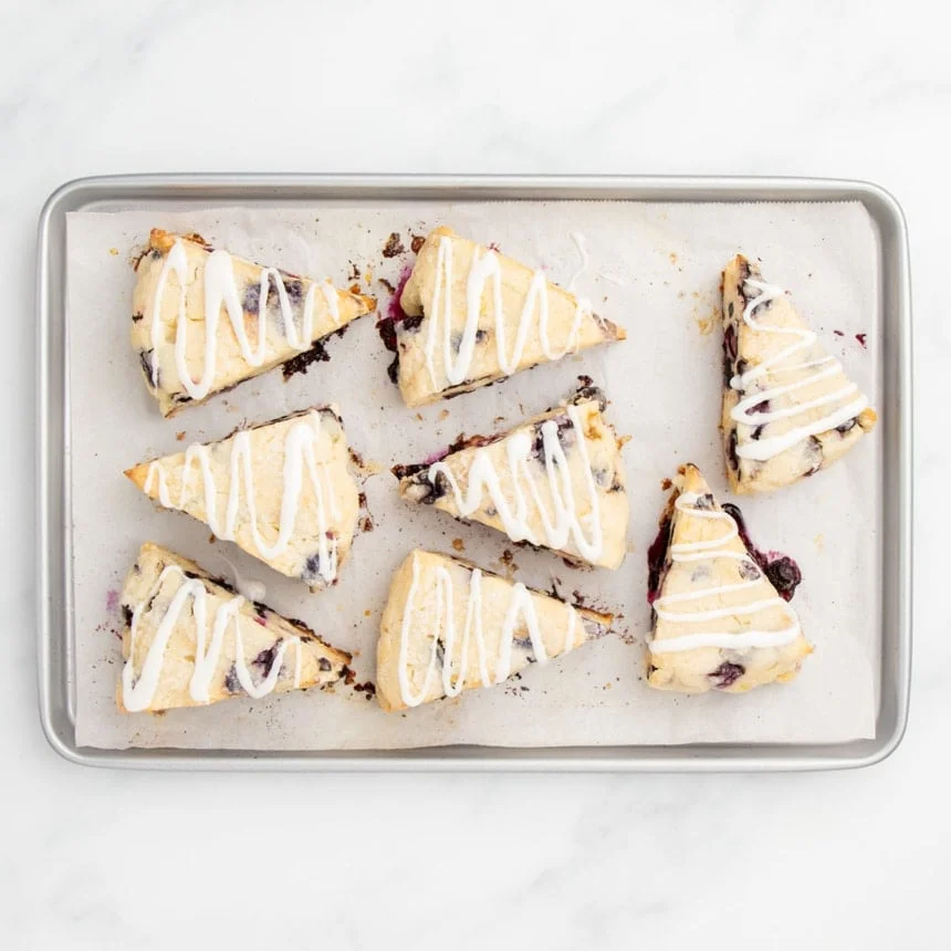 Freshly baked blueberry scones with a generous white glaze drizzled over them, on a parchment-lined baking sheet.