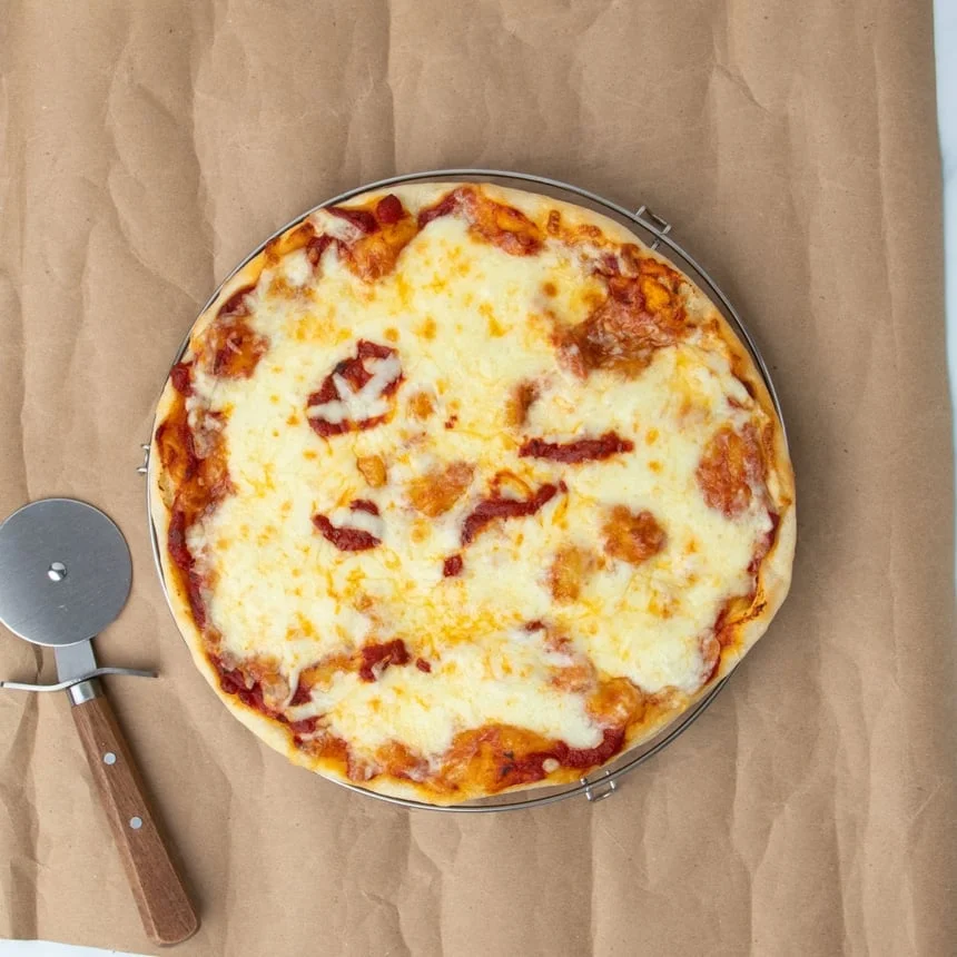 fully baked gluten free pizza round with red sauce peeking through melted off white cheese on brown paper with brown handled metal pizza wheel, all on brown kraft paper