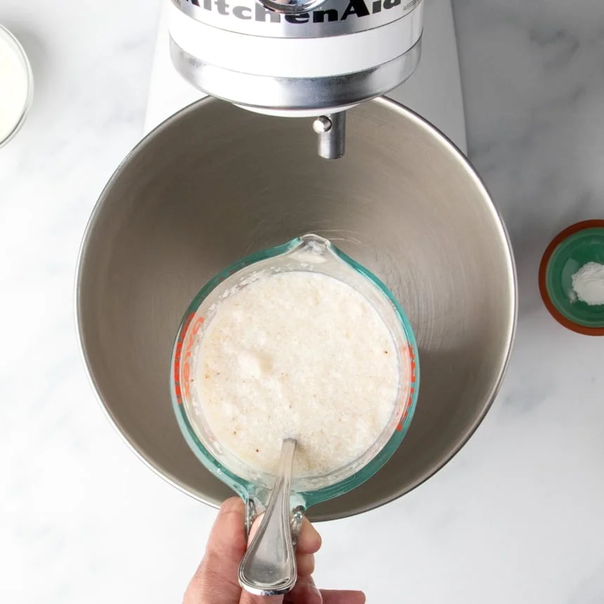 glass measuring with cloudy milk mixture with metal handle of spoon held over round metal stand mixer mixing bowl