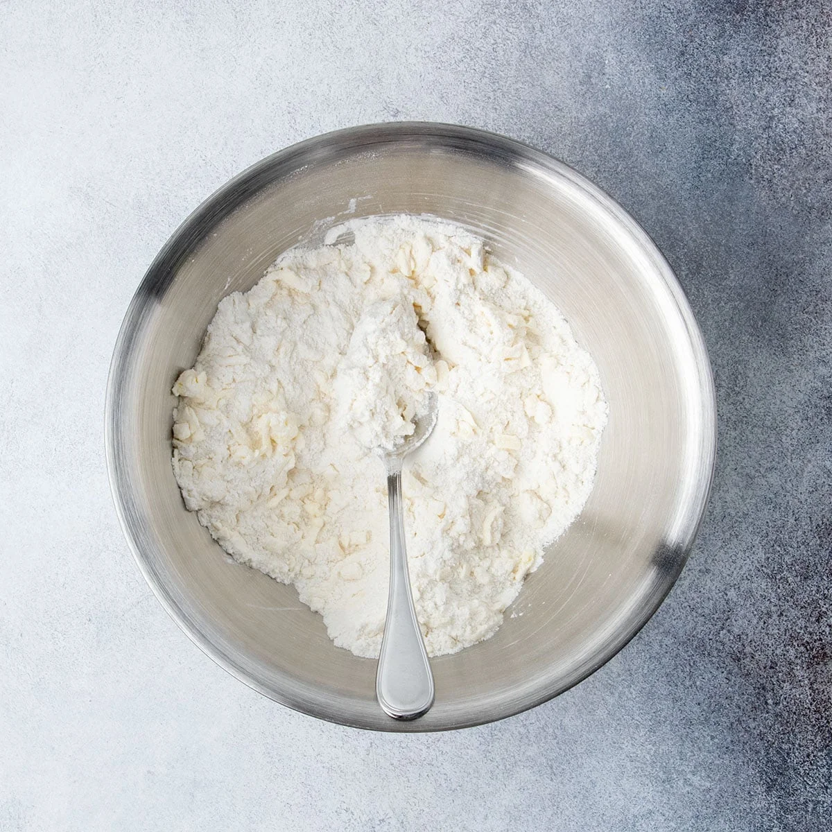 Gluten free flour and grated butter mixed together with a spoon in a metal bowl to form a loose mixture of cold grated butter in flour.