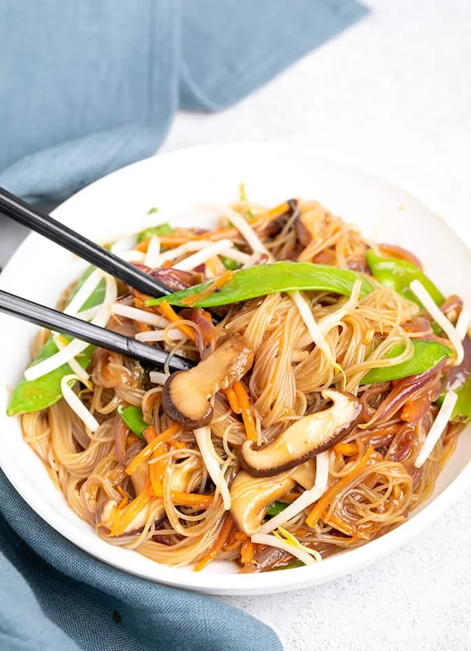 Gluten free vegetable stir fry in white bowl with black chop sticks with blue cloth