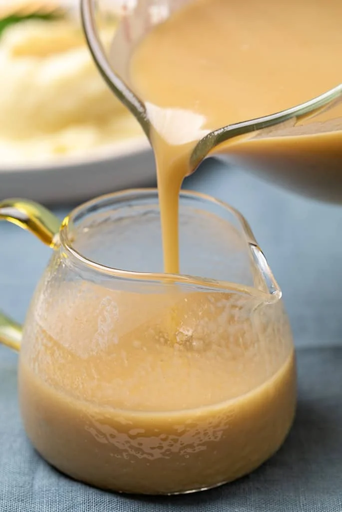 Gravy being poured from glass measuring cup into small glass server with yellow handle.