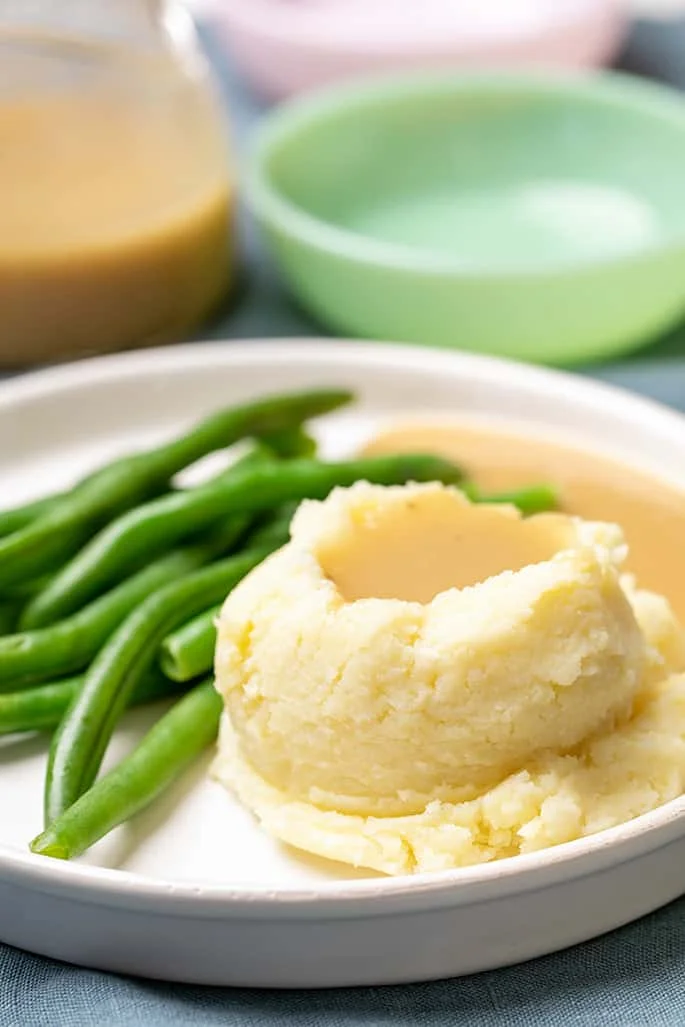 Gravy on mashed potatoes with green beans on white plate with dishes in background.