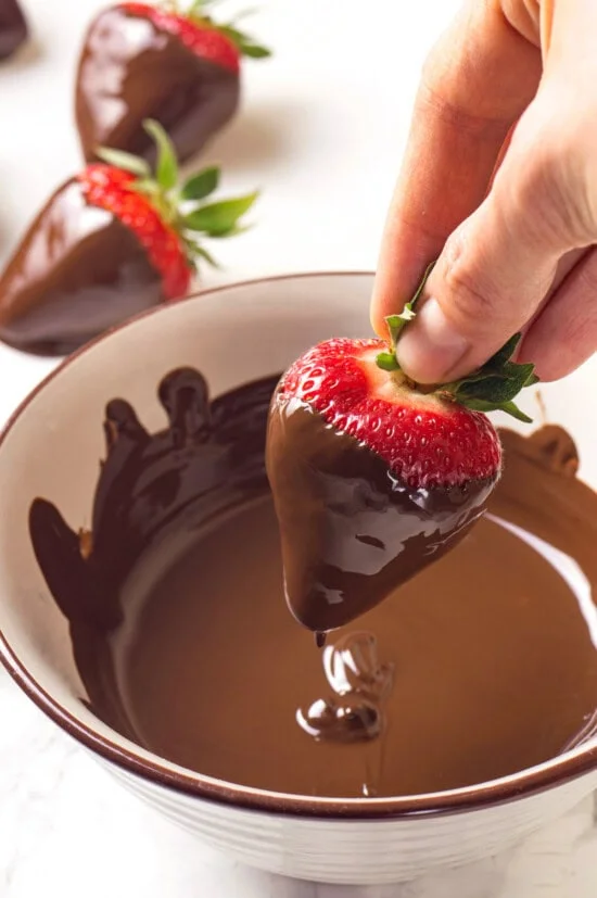 Hand dipping strawberry into bowl of melted chocolate