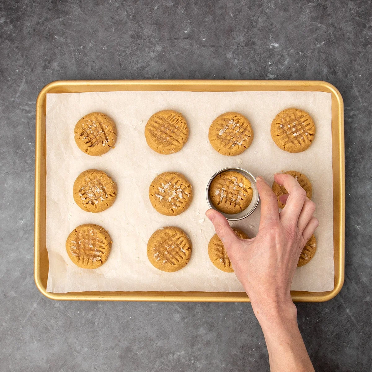 hand guiding small round cookie cutter around one of 12 freshly baked brown cookies on white paper lined gold baking sheet