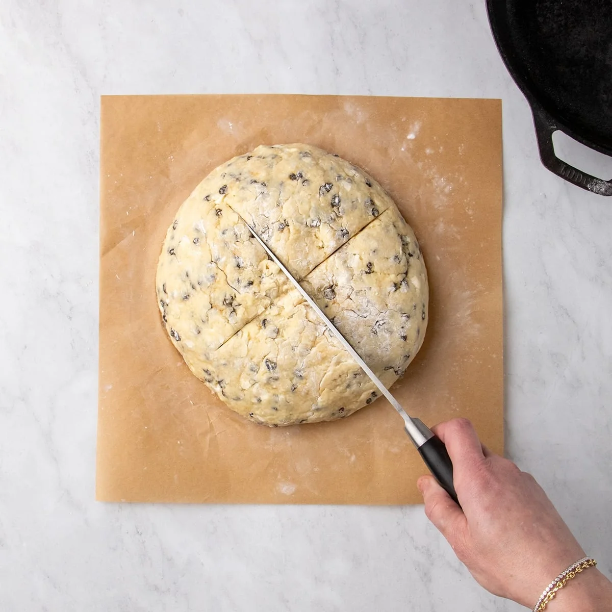 Hand holding a large metal knife with black handle cutting an X into a round of raw dough on parchment paper.