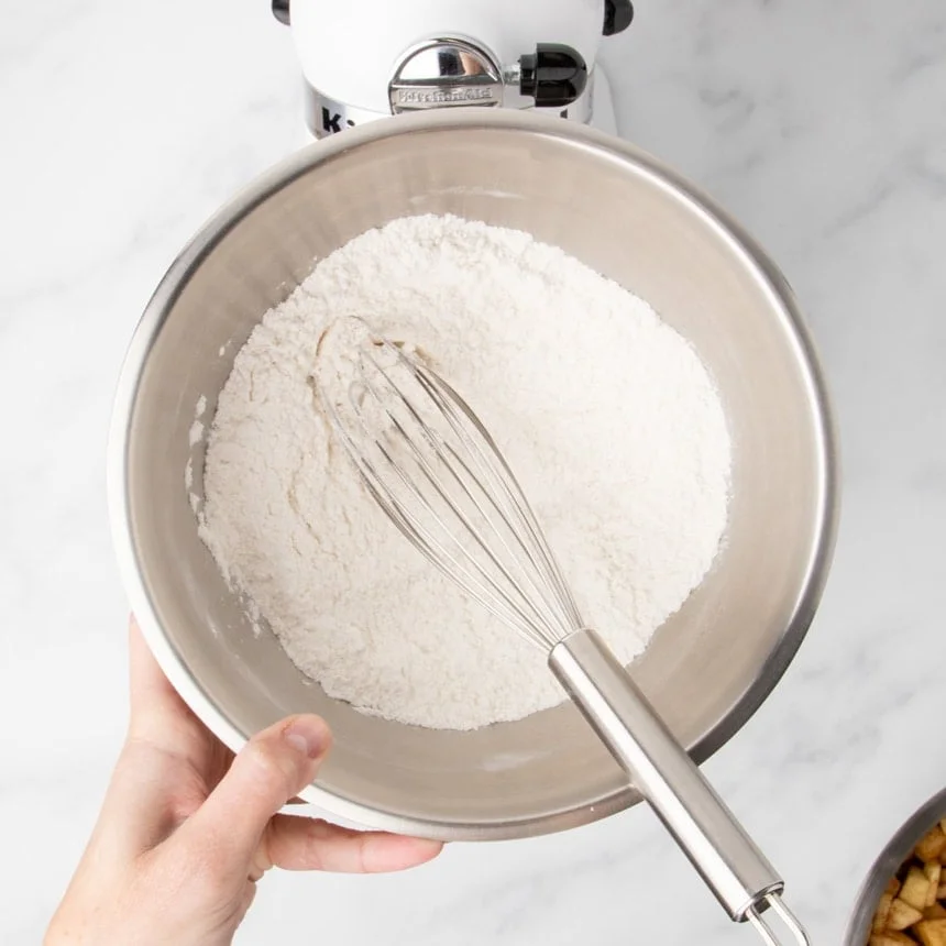 Hand holding a metal bowl of flour mixture with a whisk, mixer in the background.