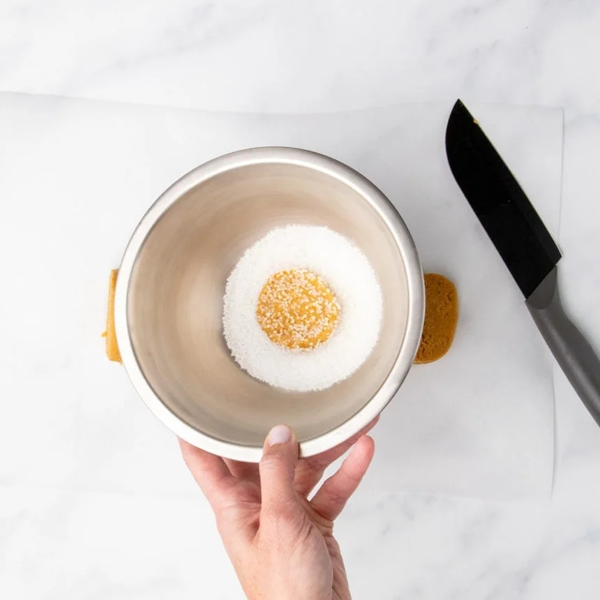 Hand holding a small stainless steel bowl with a slice of cookie dough coated in coarse sugar.