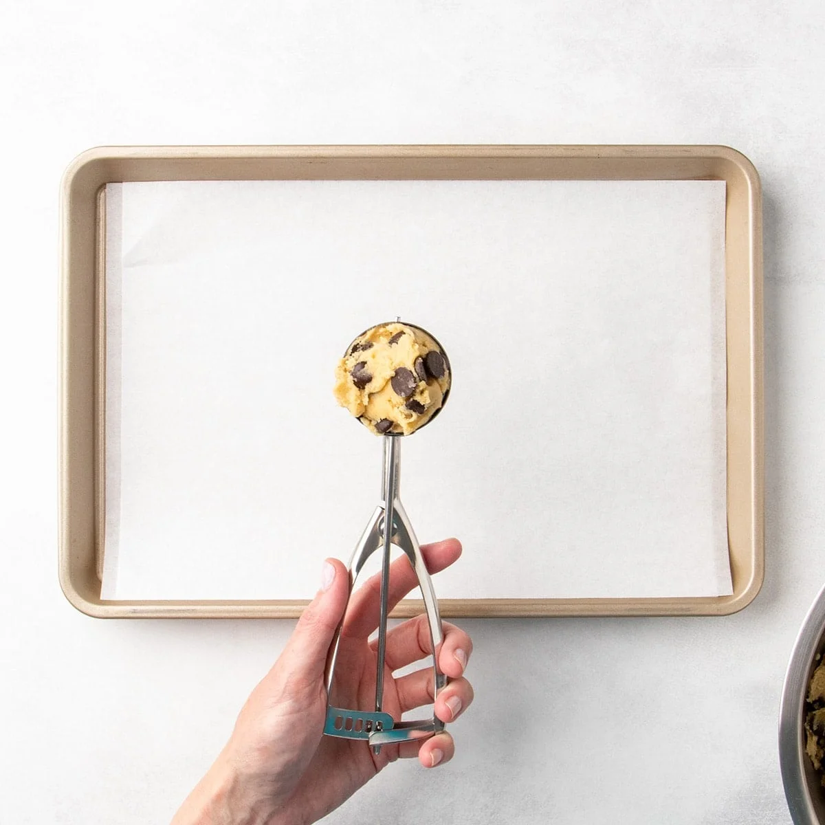 Hand holding a spring-loaded cookie scoop with chocolate chip cookie dough above a parchment-lined baking sheet.