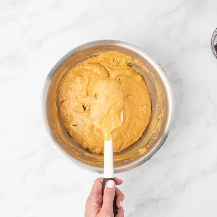 Hand holding a white spatula with thick, light orange raw muffin batter above the mixing bowl with the rest of the batter.