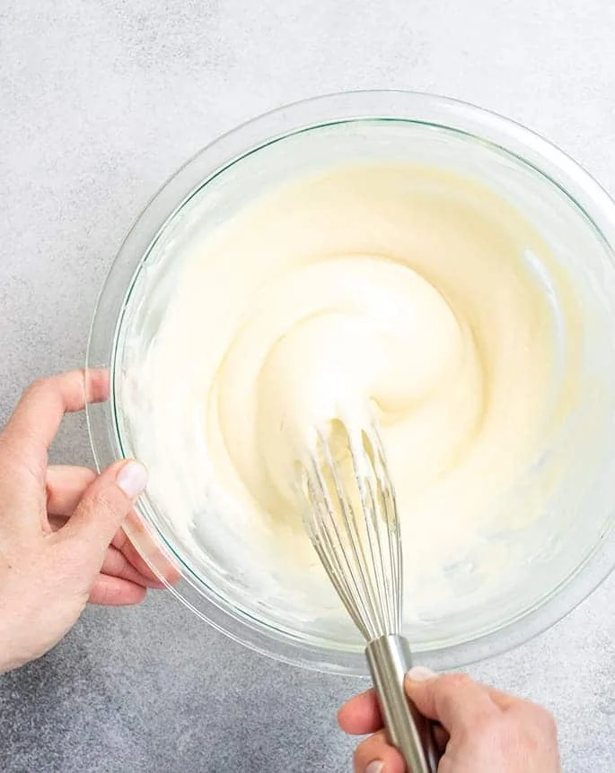 Hand holding glass bowl of pancake batter and hand holding whisk mixing the batter