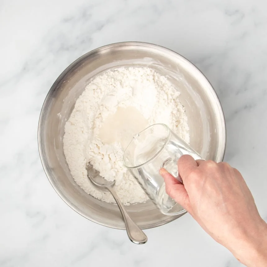 hand holding glass measuring cup pouring water into mixing bowl with large metal mixing spoon in bowl