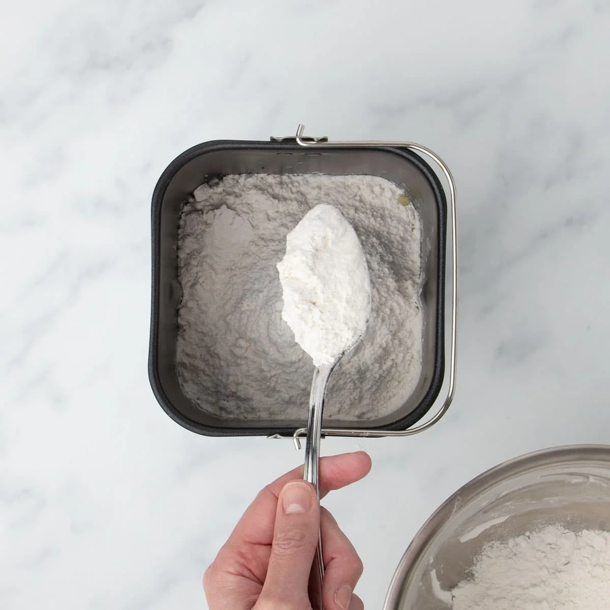 hand holding large metal mixing spoon filled with white flour mixture turning flour over into black bread machine baking pan with more flour