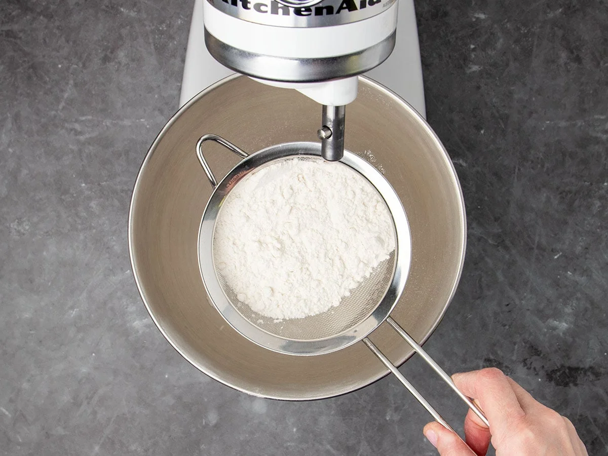 hand holding medium-size metal sieve with white flour over mixing bowl