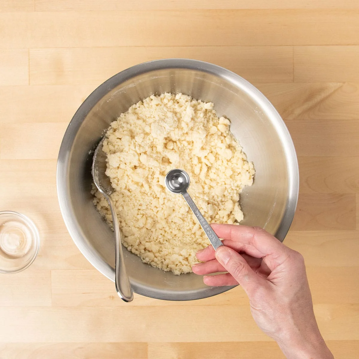 hand holding metal half teaspoonful of water over crumbly raw light yellow cookie mixture in same mixing bowl