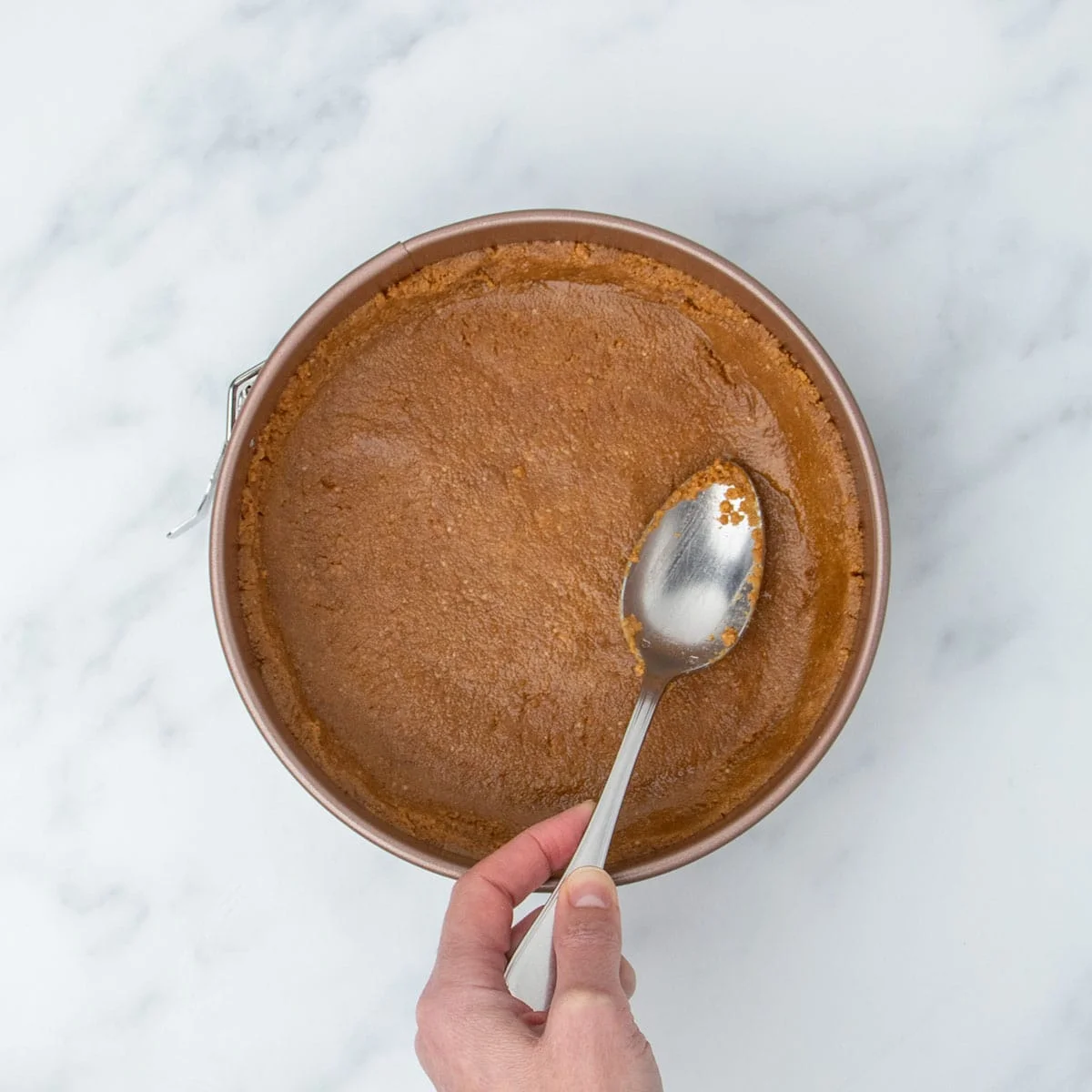 hand holding metal mixing spoon pressing brown graham cracker crust into even layer in gold spring form pan